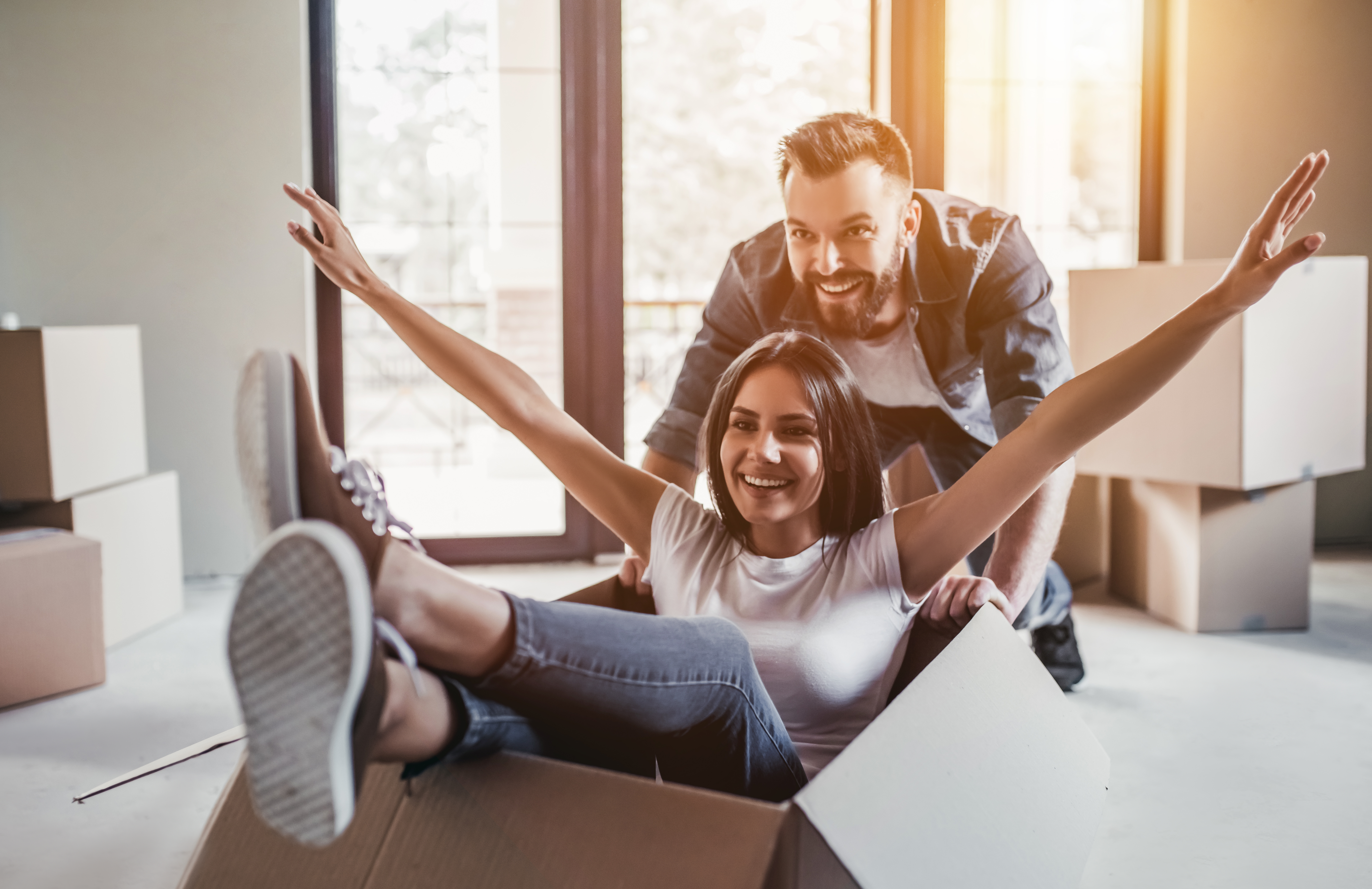 An image of a girl sitting happily in a box used for moving in and her husband/boyfriend dragging the box